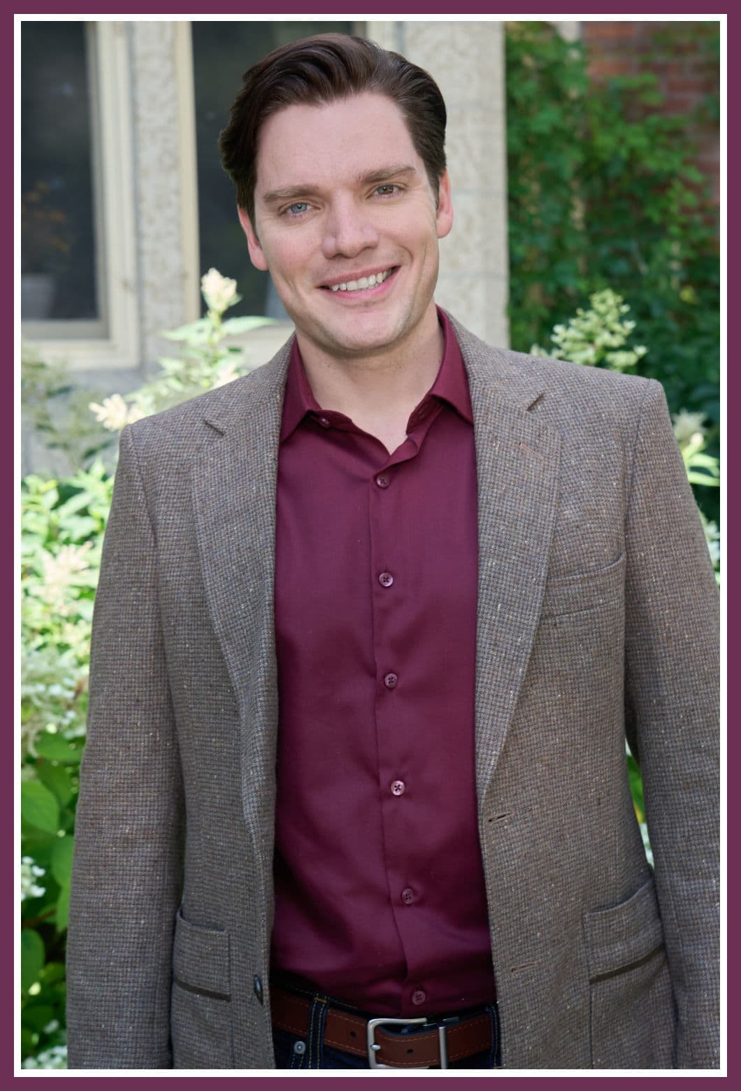 Dominic Sherwood in character as Brian/Malcolm from the Hallmark movie Haunted Wedding, wearing a maroon shirt and tweed jacket, smiling outdoors.